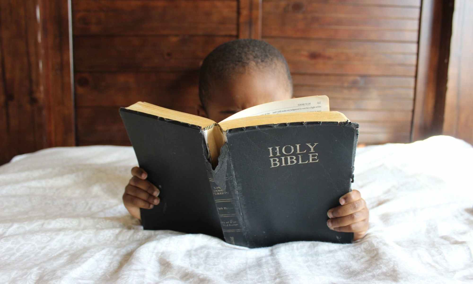 Black boy reading holy bible under white covers in bed