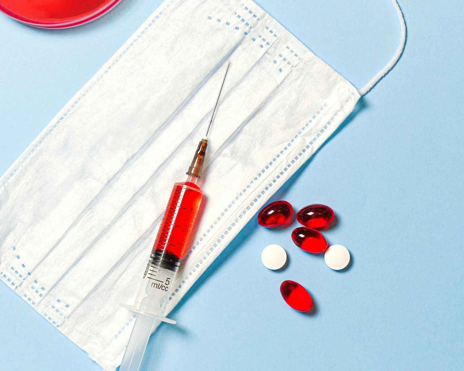 Syringe, red and white pills next to face mask on blue background