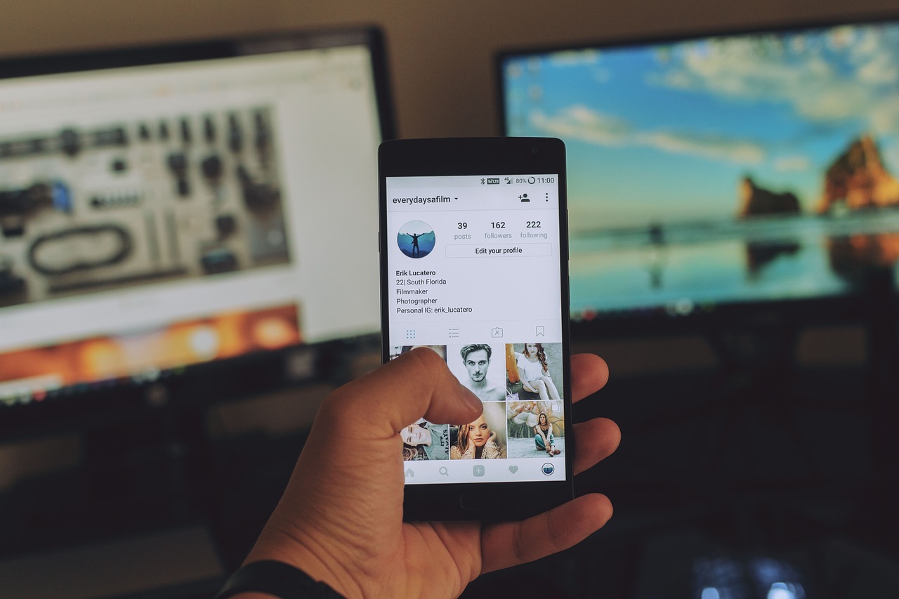 Hand holding phone with Instagram open near two flat computer monitors